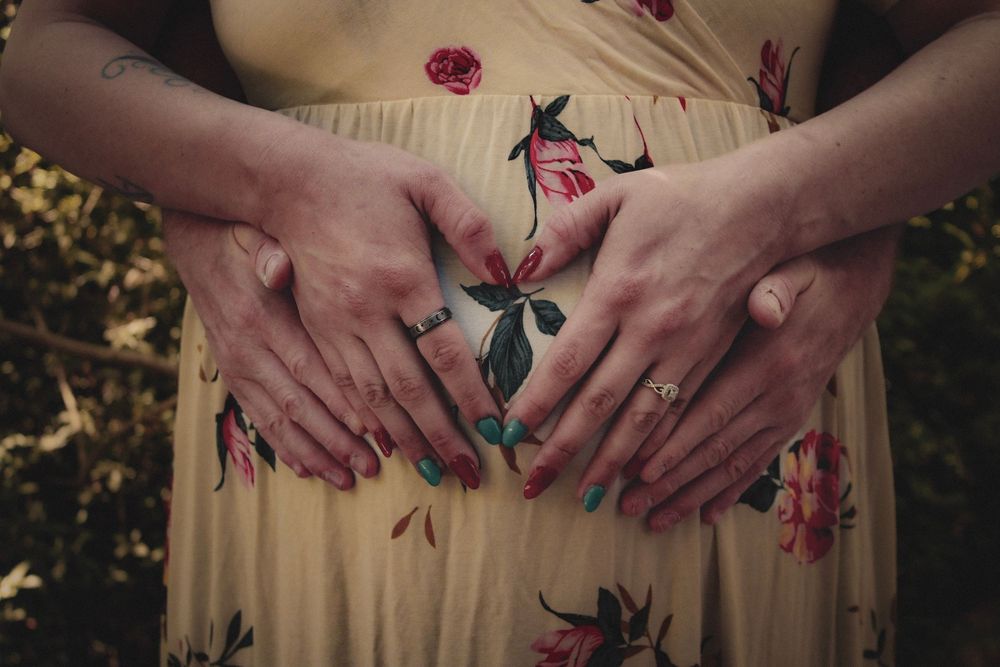 Two pairs of hands forming a heart shape on a pregnant woman's belly in a floral yellow dress.