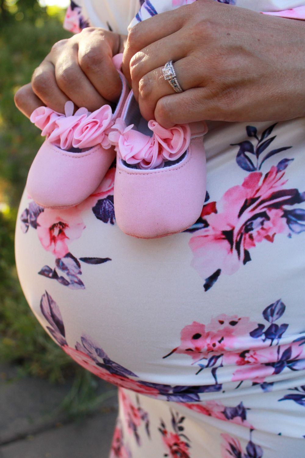 Pregnant woman in floral dress holding tiny pink baby girl shoes with rose details over her baby bump.