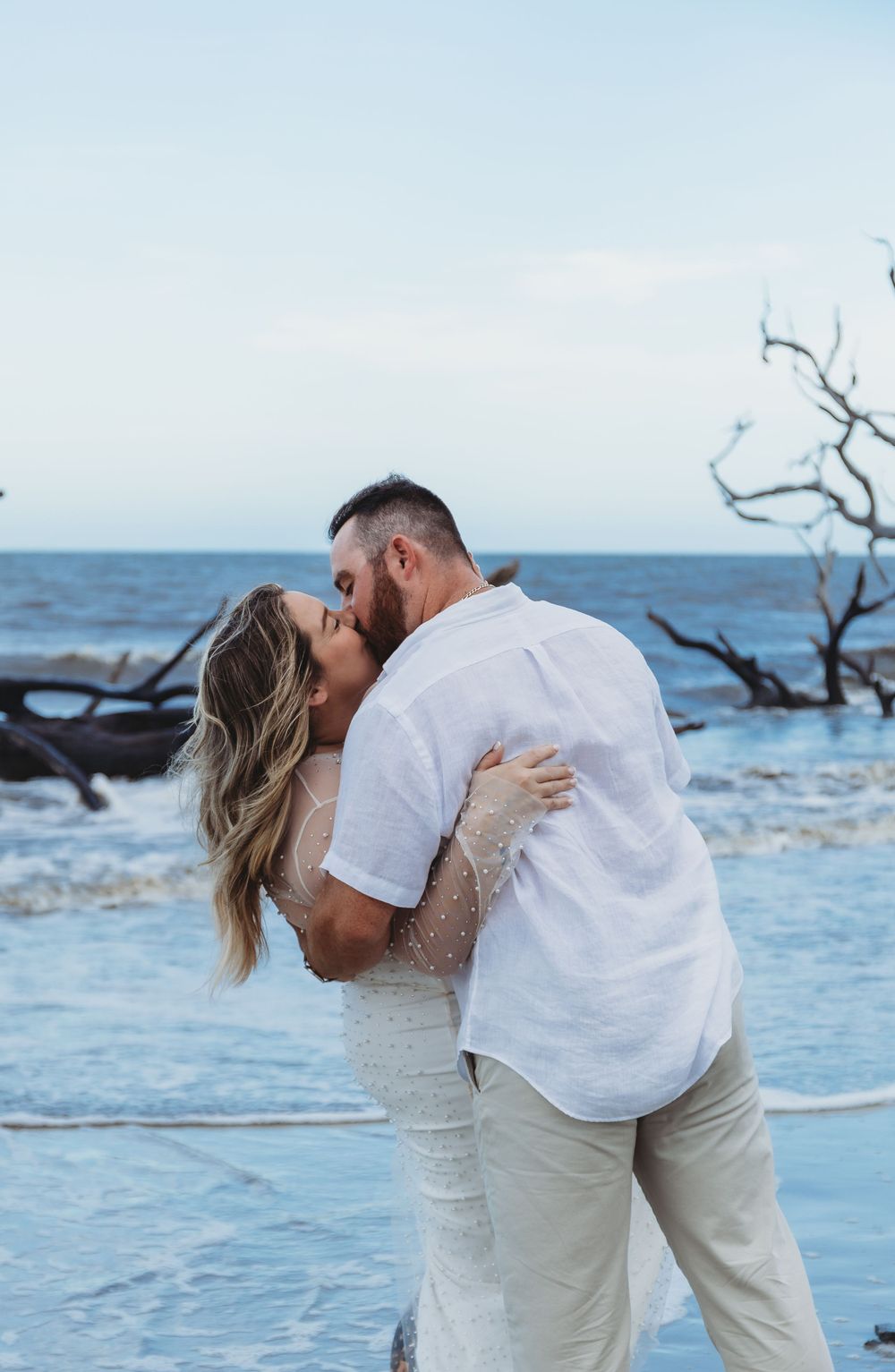 A couple shares a passionate kiss on the beach with driftwood and waves in the background.