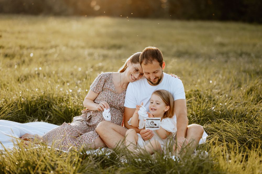 Babybauchshooting von Familie, die in einer Wiese sitzt und Babyschuhe und Ultraschallbild zeigt Mengen Jovanna Burkard