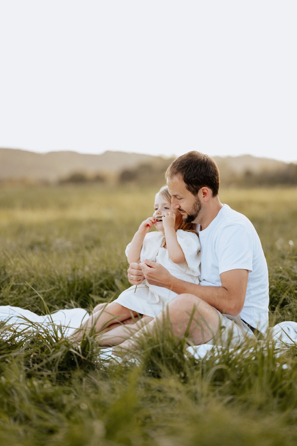 Familienbild von Babybauchshooting von Vater, der mit Tochter auf einer Wiese sitzend kuschelt Mengen Jovanna Burkard