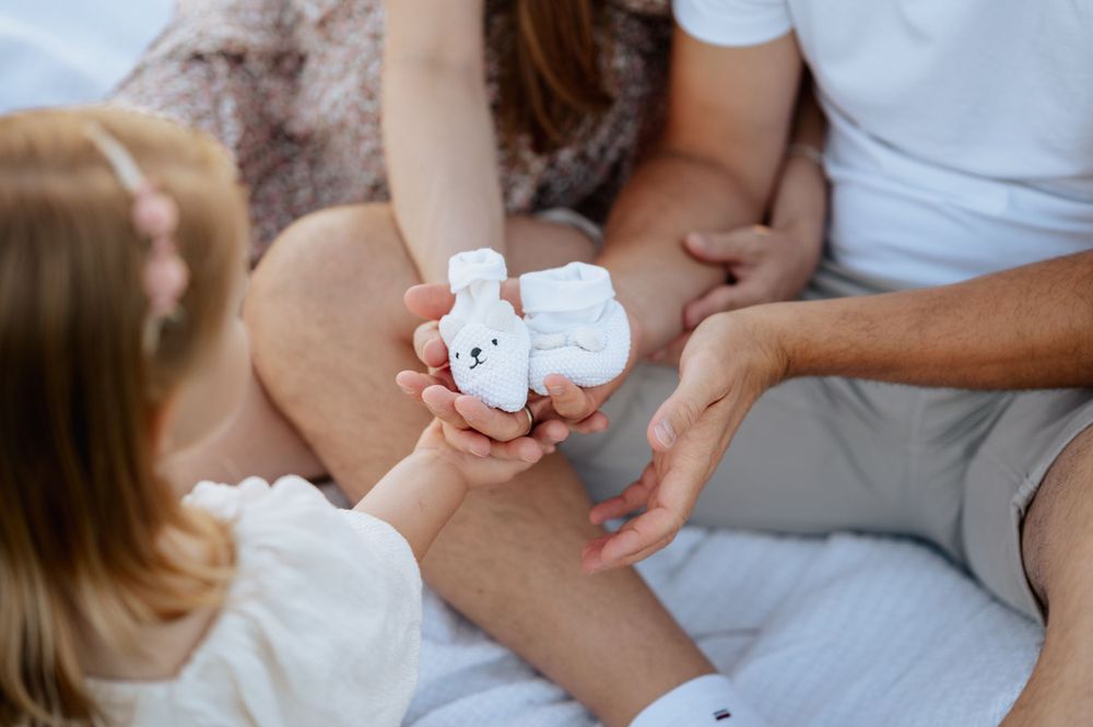Babybauchshooting von Familie, die auf einer Decke sitzen und Babyschuhe in den Händen halten Mengen Jovanna Burkard