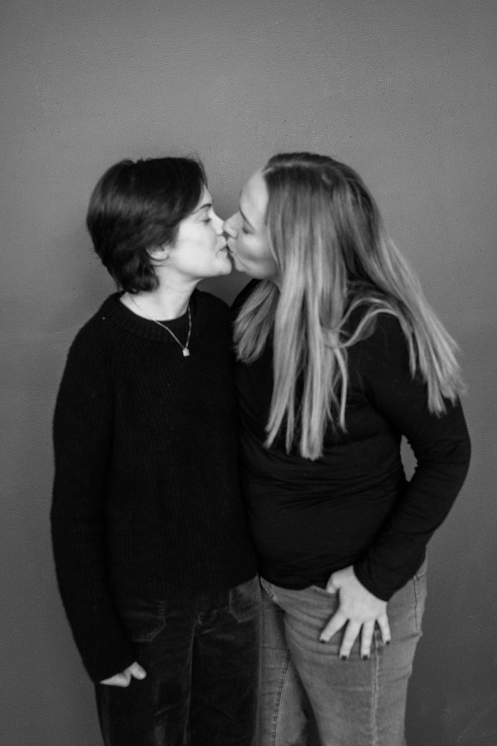 Black and white portrait of two people in dark clothing sharing an intimate kiss against a plain background.