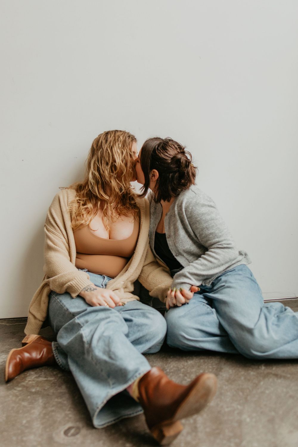 Two people in casual clothing sit intimately together on a concrete floor in a minimalist setting.