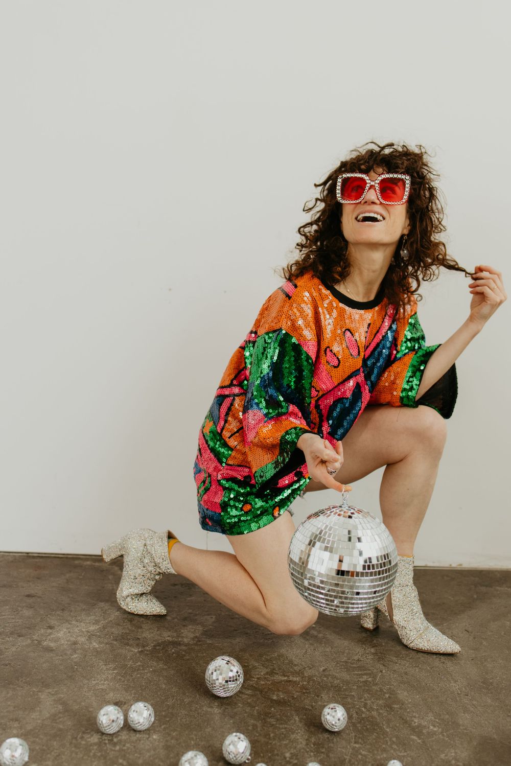Retro fashion shoot with disco balls featuring colorful sequin outfit and white boots on concrete floor.
