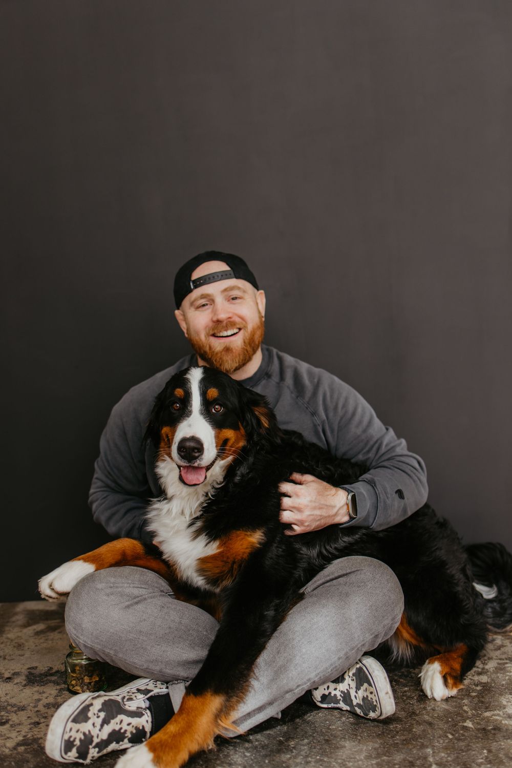 A Bernese Mountain Dog sits happily on the ground with someone in a grey sweatshirt and baseball cap.