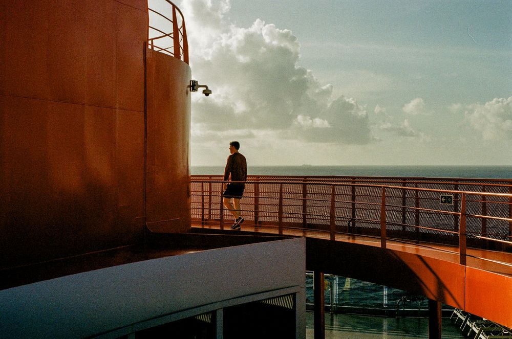 A silhouetted figure stands on a cruise ship deck at sunset against an orange and blue sky.