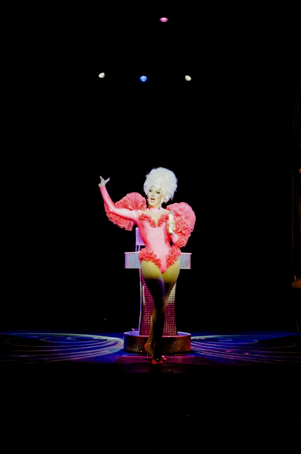 A performer in a sparkly pink costume on stage under dramatic spotlights.