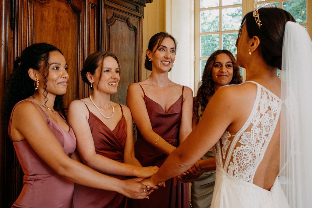 Bride in lace-back gown holding hands with four smiling bridesmaids in mauve satin dresses indoors.