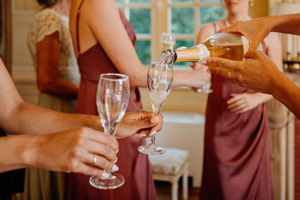 Champagne being poured into flutes held by bridesmaids in dusty rose dresses during wedding preparations.