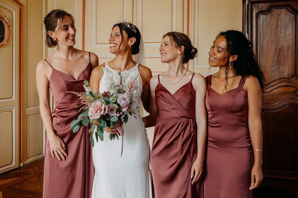 Bride in white lace gown holding bouquet laughing with three bridesmaids in dusty rose satin dresses.