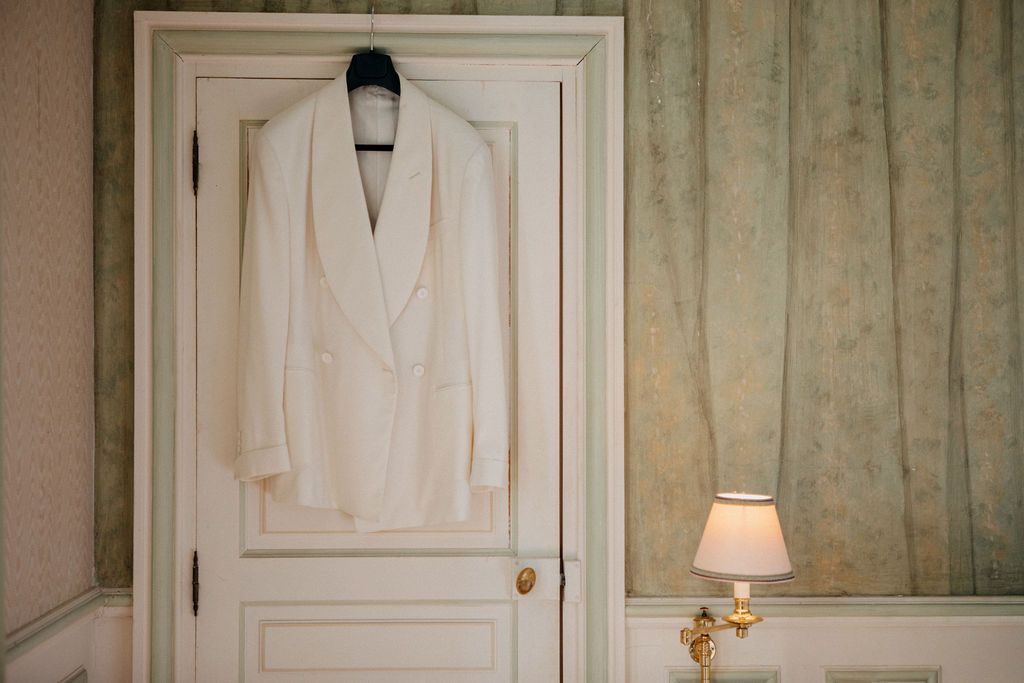 White double-breasted groom's tuxedo jacket hanging on door in elegant room with gold lamp.
