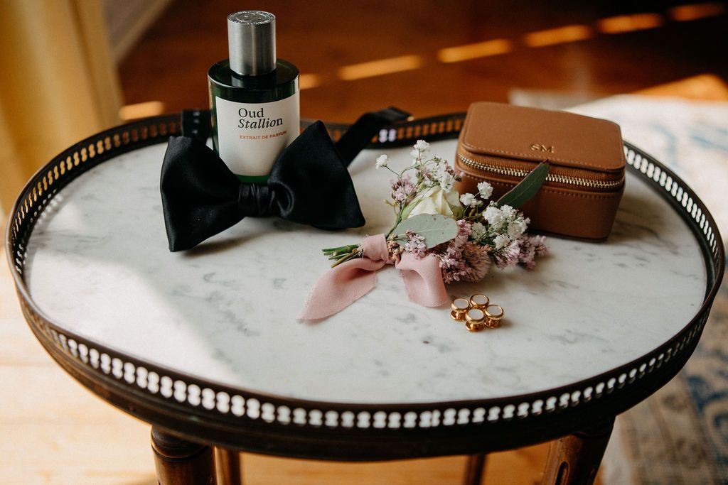 Wedding flat lay with Oud Satin perfume, black bow tie, floral boutonniere, and tan jewelry box.