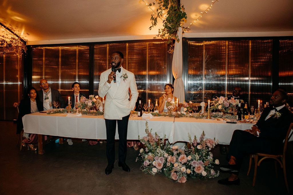 Groom in white tuxedo giving a speech at wedding reception with floral arrangements on head table.