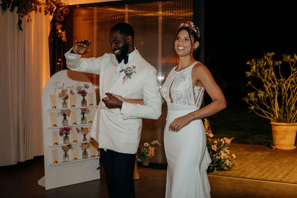 Bride and groom smiling beside a floral seating chart display at their outdoor wedding reception.
