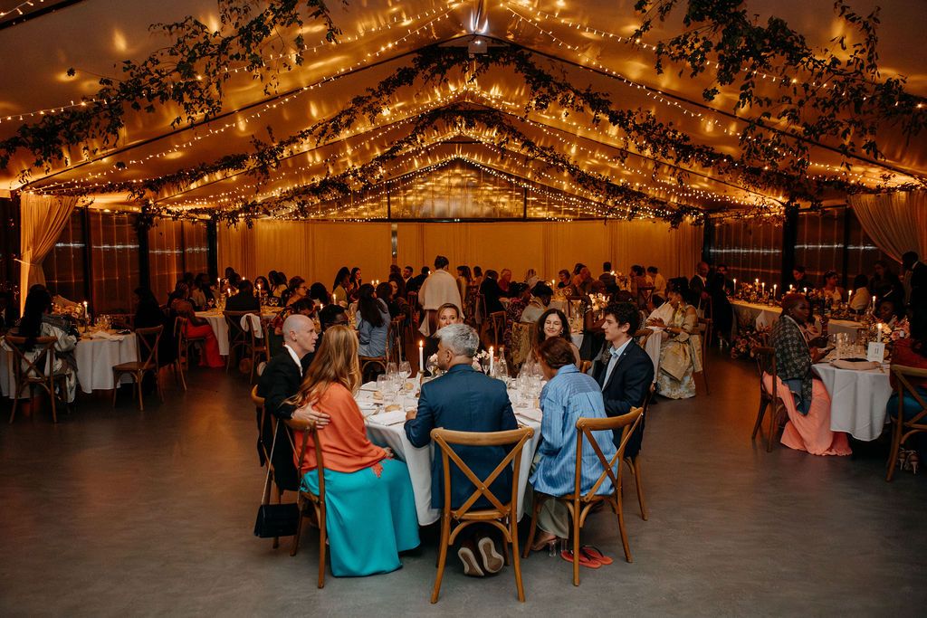Wedding reception venue filled with guests dining under a canopy of warm fairy lights and greenery.