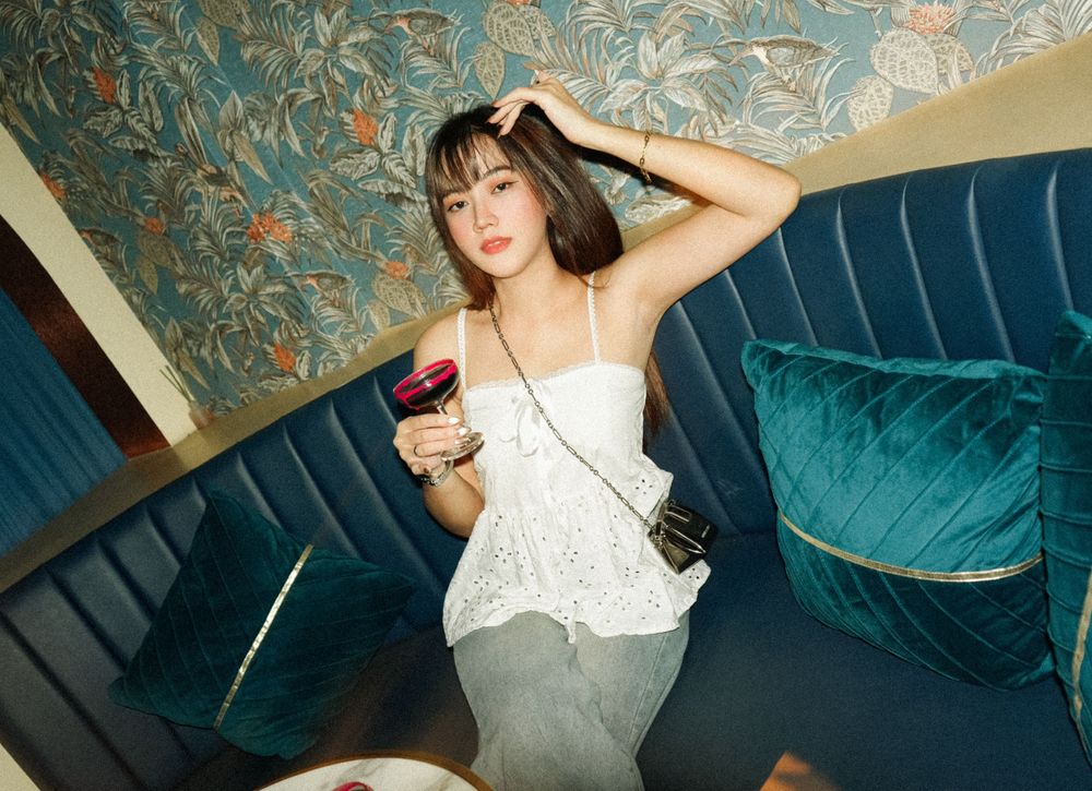 A person in white lounges on a teal velvet couch against a floral wallpapered wall.