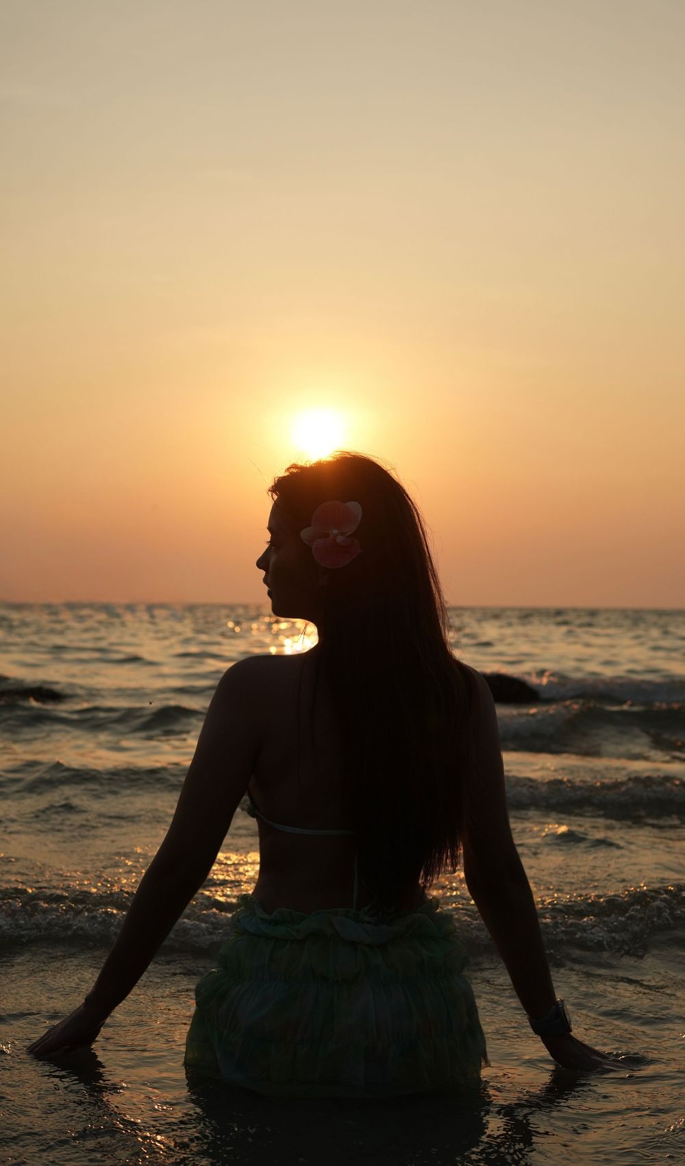 Silhouette of a person sitting in shallow ocean water at sunset with golden light reflecting on the waves.