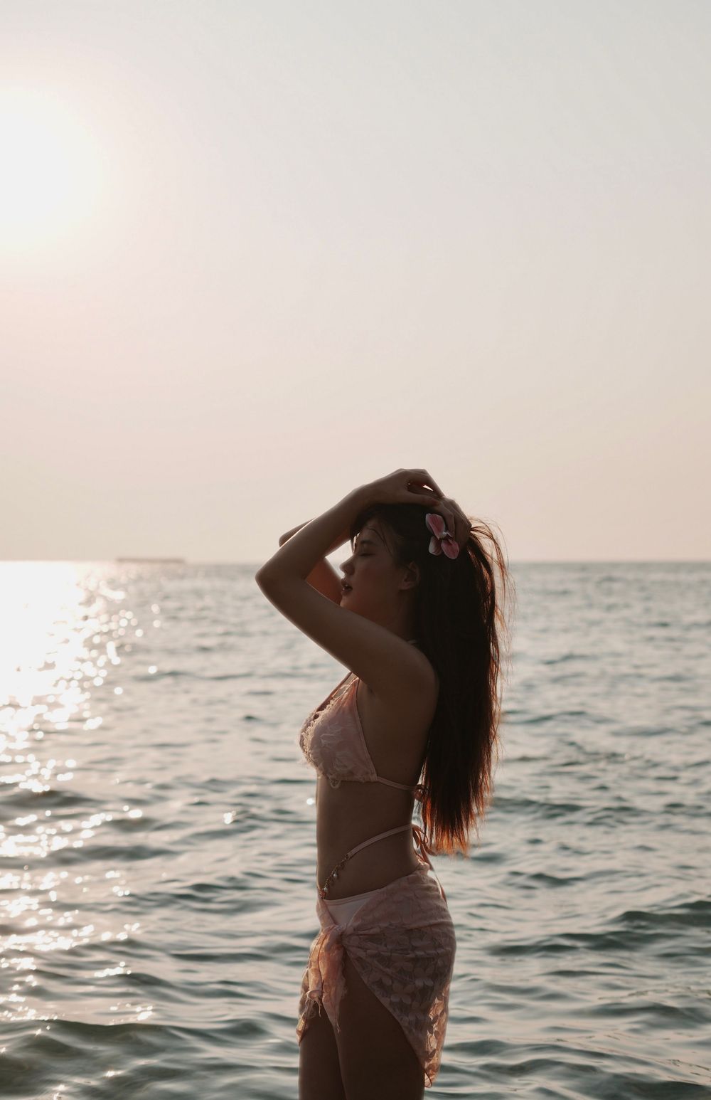 Woman in white dress poses gracefully against ocean backdrop during sunset in artistic beach photoshoot series.
