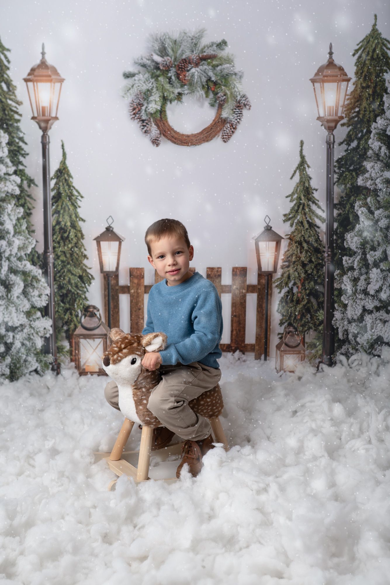 Winter holiday scene with a young child and teddy bear sitting in a snowy setting with glowing lanterns and evergreen decor.