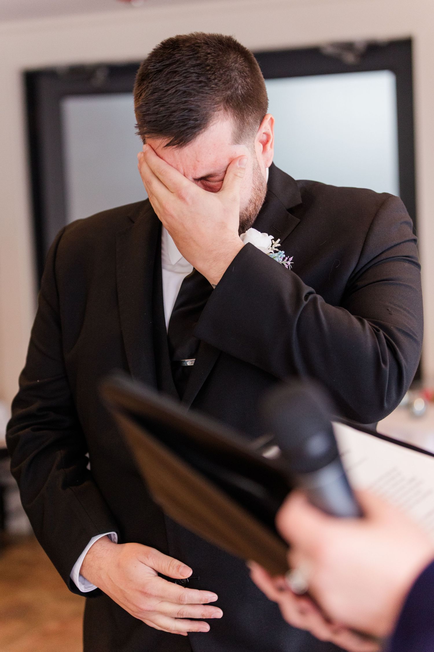 Emotional groom wipes tears from his eyes during wedding ceremony while wearing black suit.