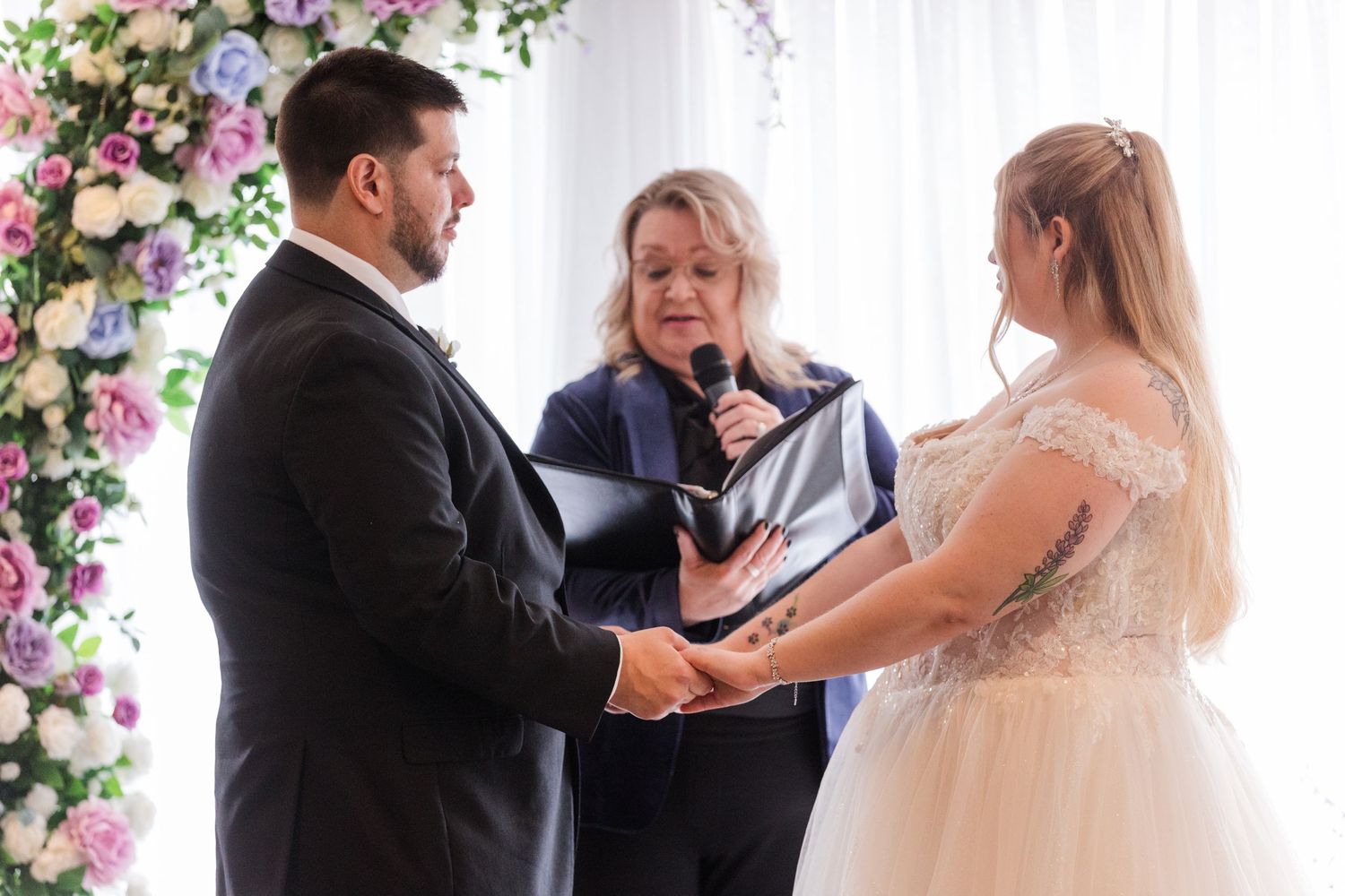 A wedding ceremony takes place under a floral arch with pink and purple blooms.
