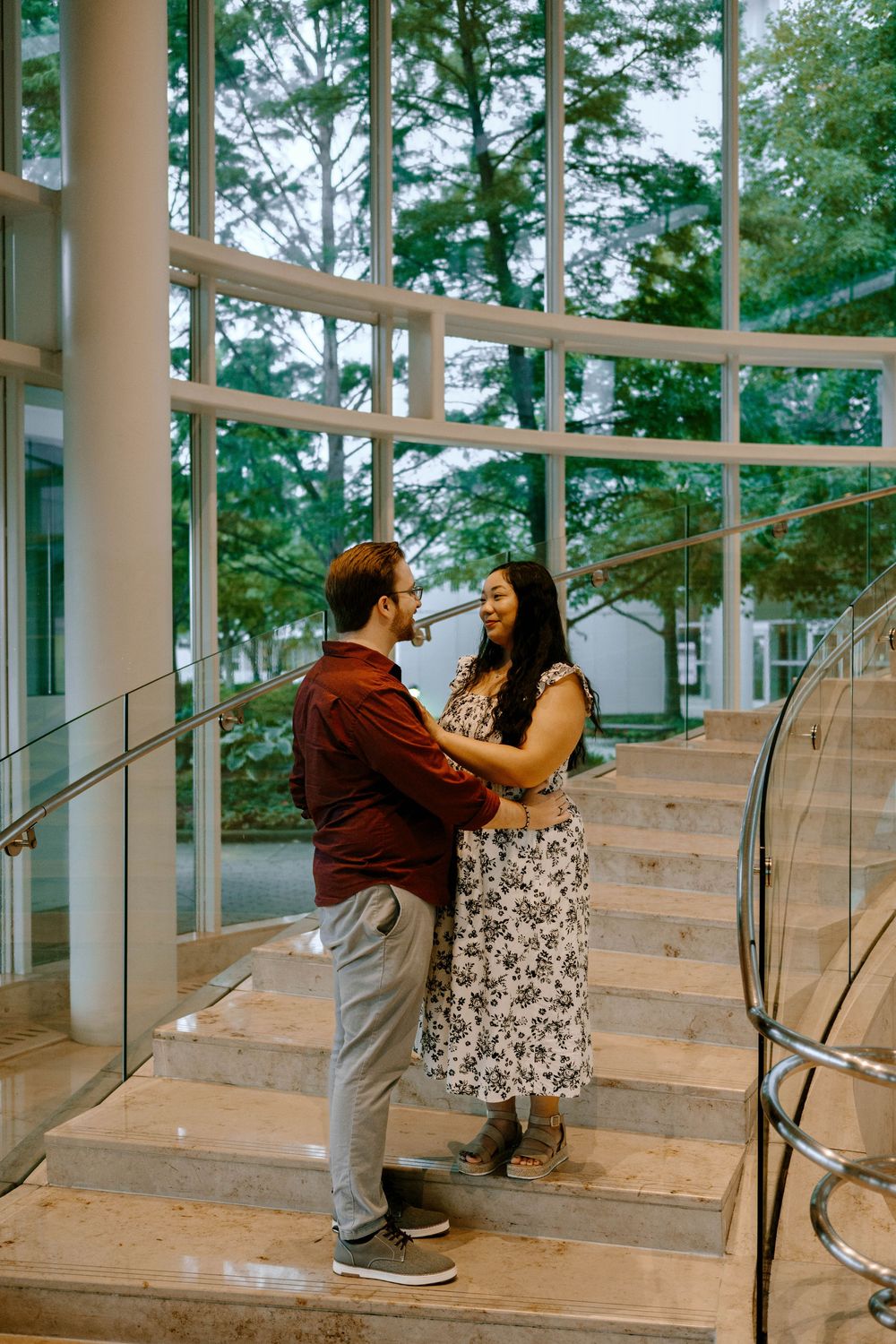 A couple shares a romantic moment on wooden steps surrounded by glass walls and greenery in an outdoor setting.