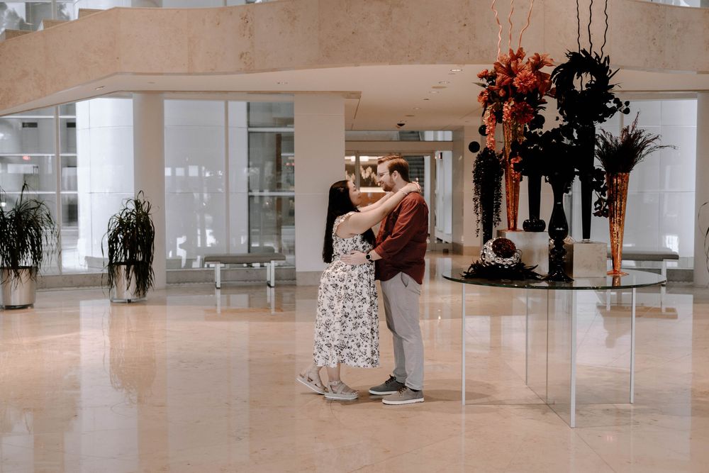 A couple embraces in an elegant lobby space decorated with tall floral arrangements and white columns.