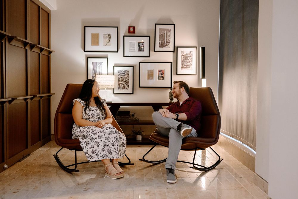 Two people sit in modern rocking chairs having a conversation in a corner decorated with framed artwork.