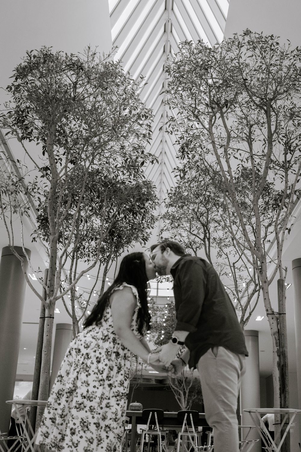A romantic couple shares a tender moment in a modern indoor space with tall potted trees and white columns.