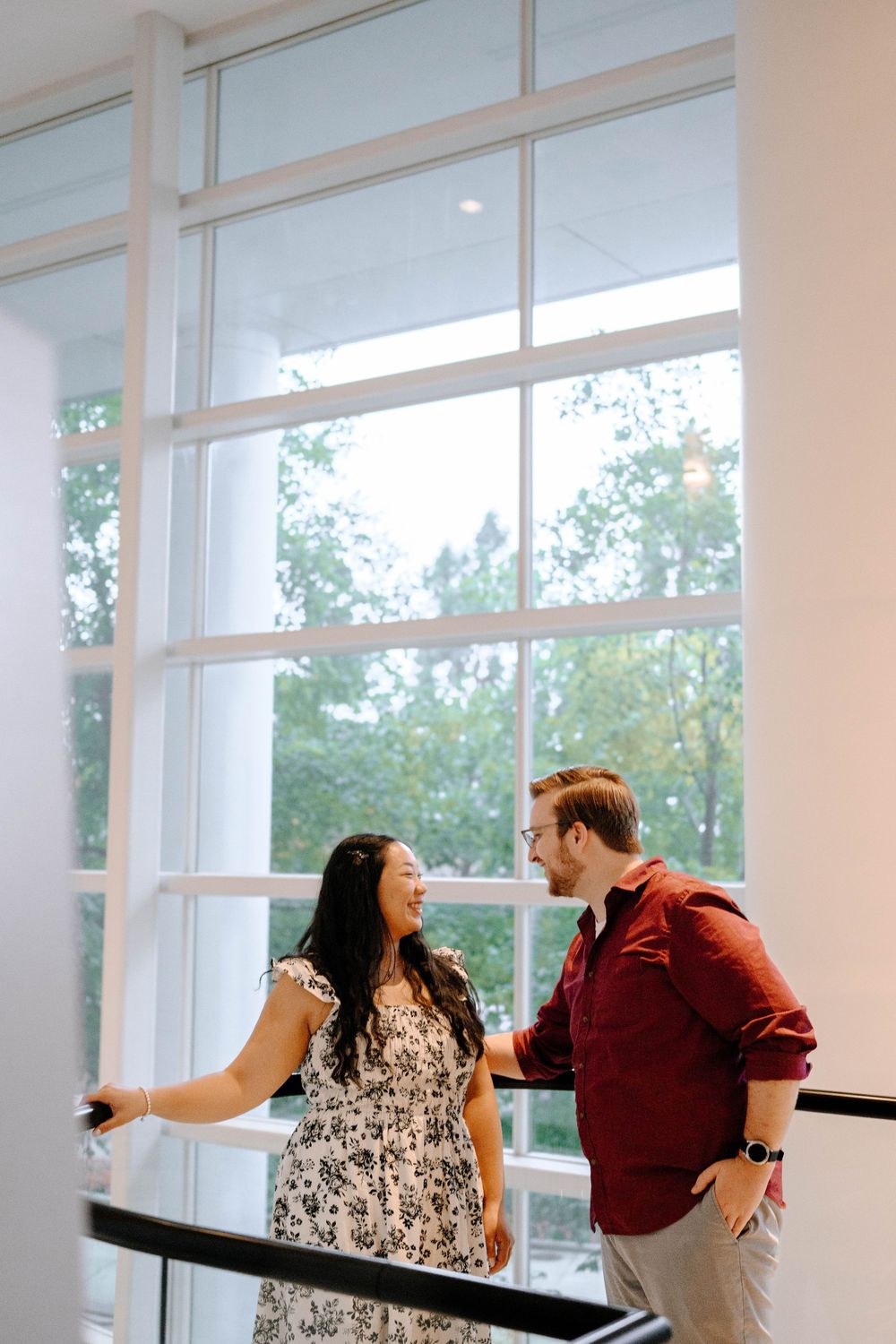 A couple in casual attire share a playful moment near large windows in a bright indoor space.