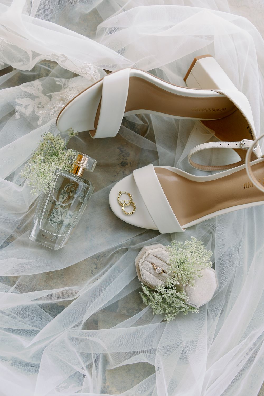 Elegant white bridal shoes with veil and perfume bottle on light fabric.