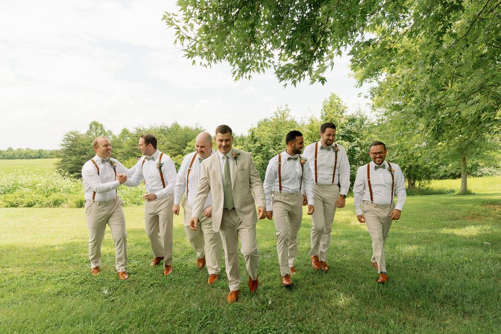 Wedding party in light colored attire poses together on a lush green lawn.