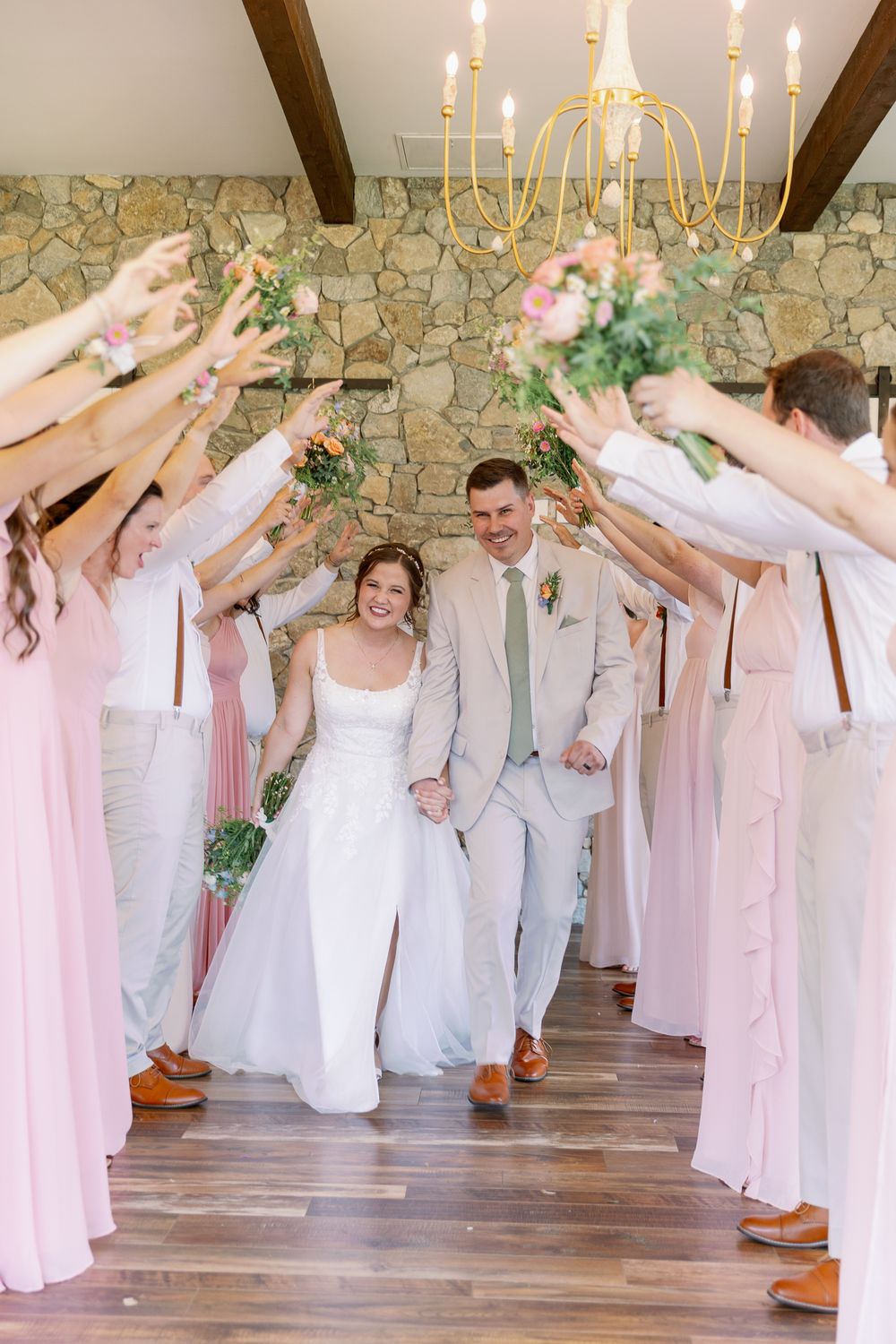 Wedding party in pink dresses and light suits creating flower arch tunnel for newlyweds.