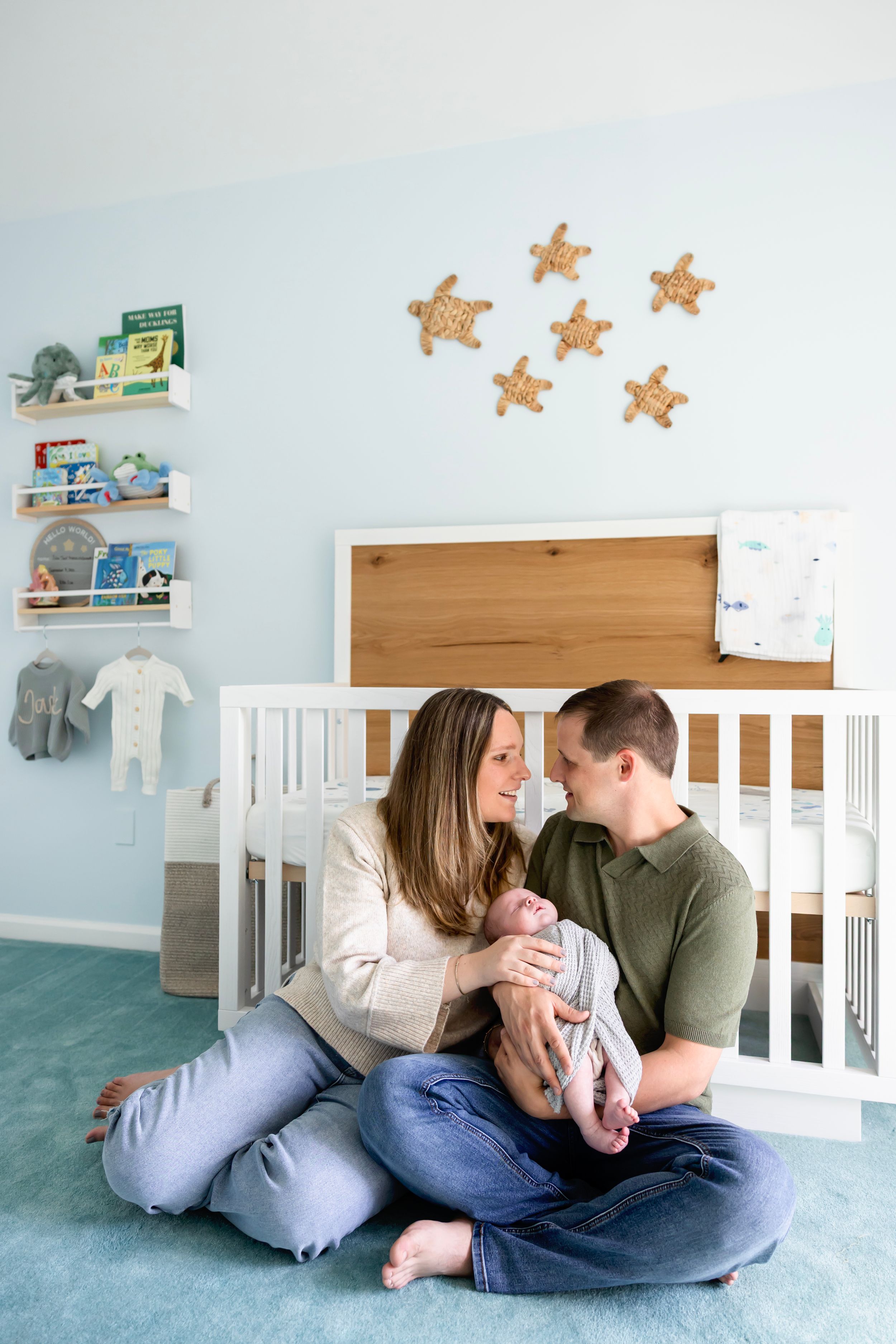 A couple sits on a nursery floor with their newborn baby, with star decorations and shelves visible on the blue walls.