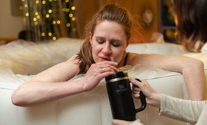 Someone relaxes on a couch with a warm beverage in a mug during the holiday season with Christmas lights in background.