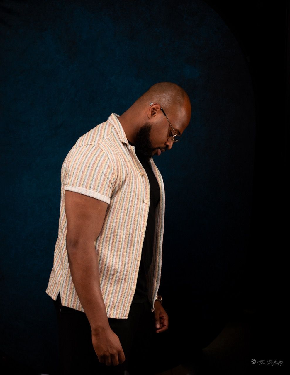Bald man with beard in striped short-sleeve shirt looking down against dark blue background.