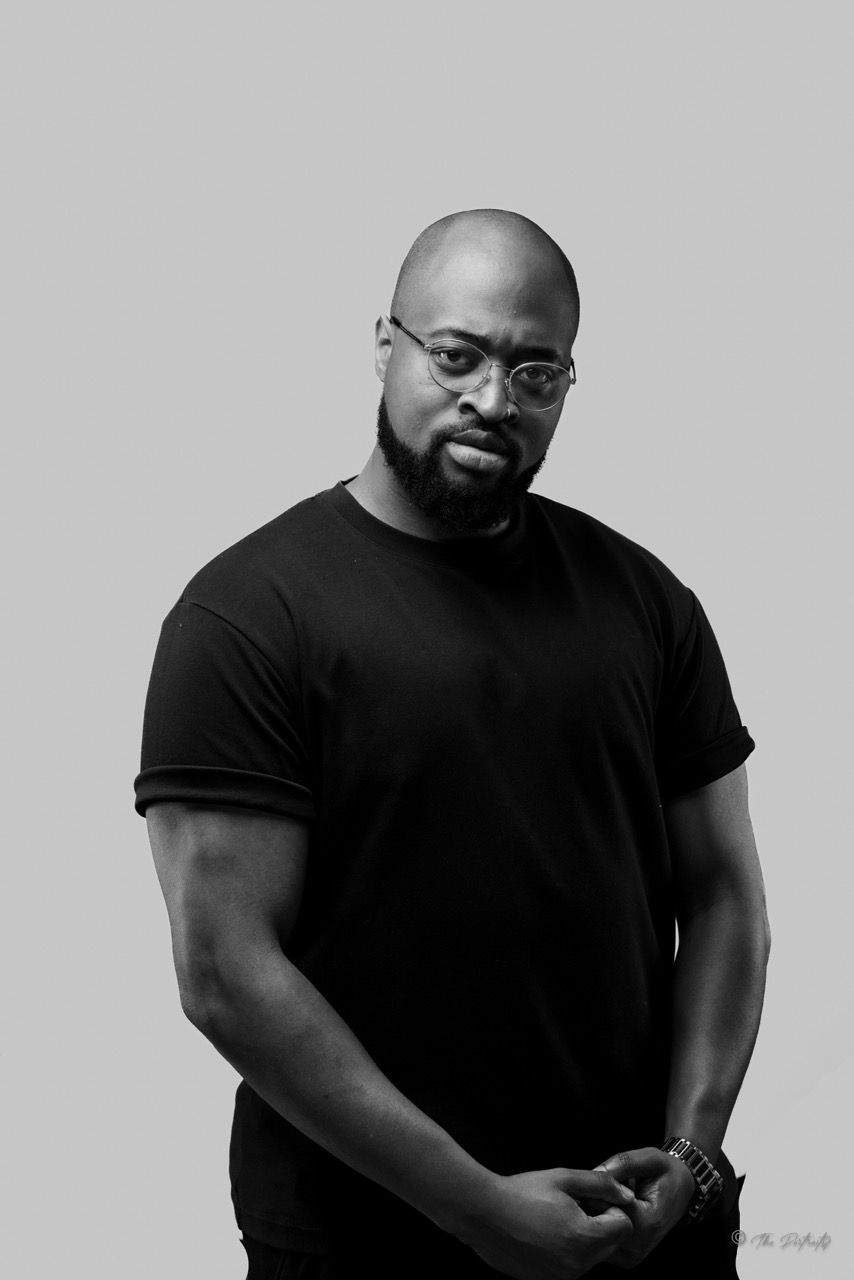 Muscular bald man with beard wearing black t-shirt and glasses in black and white studio portrait.