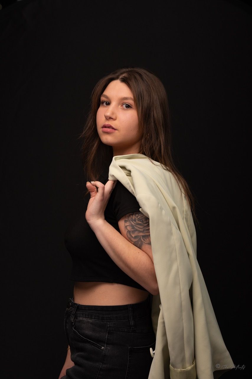 Young woman with long brown hair and arm tattoo holding white fabric over shoulder on black background.
