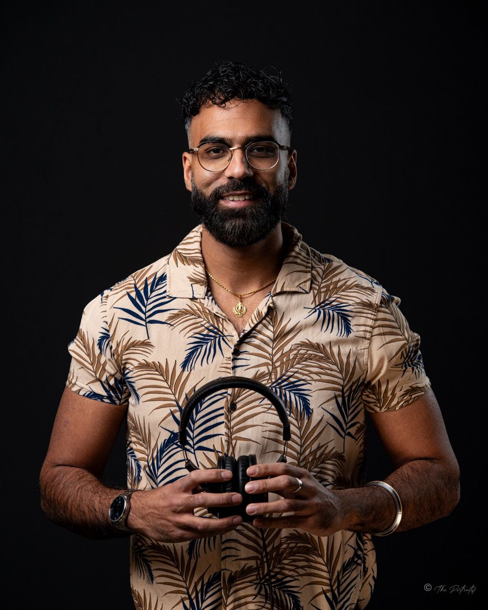 Smiling bearded man in tropical floral shirt holding DJ headphones in studio portrait.