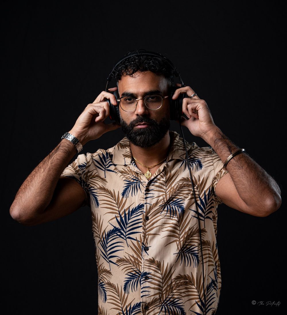 Bearded man in tropical print shirt adjusting headphones against dark background.