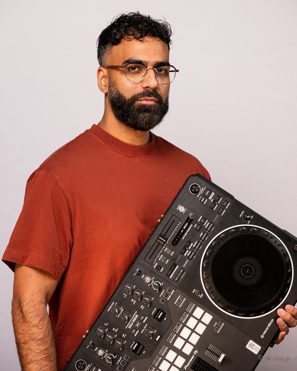 Bearded man in rust red t-shirt holding Pioneer DJ mixer controller in studio portrait.