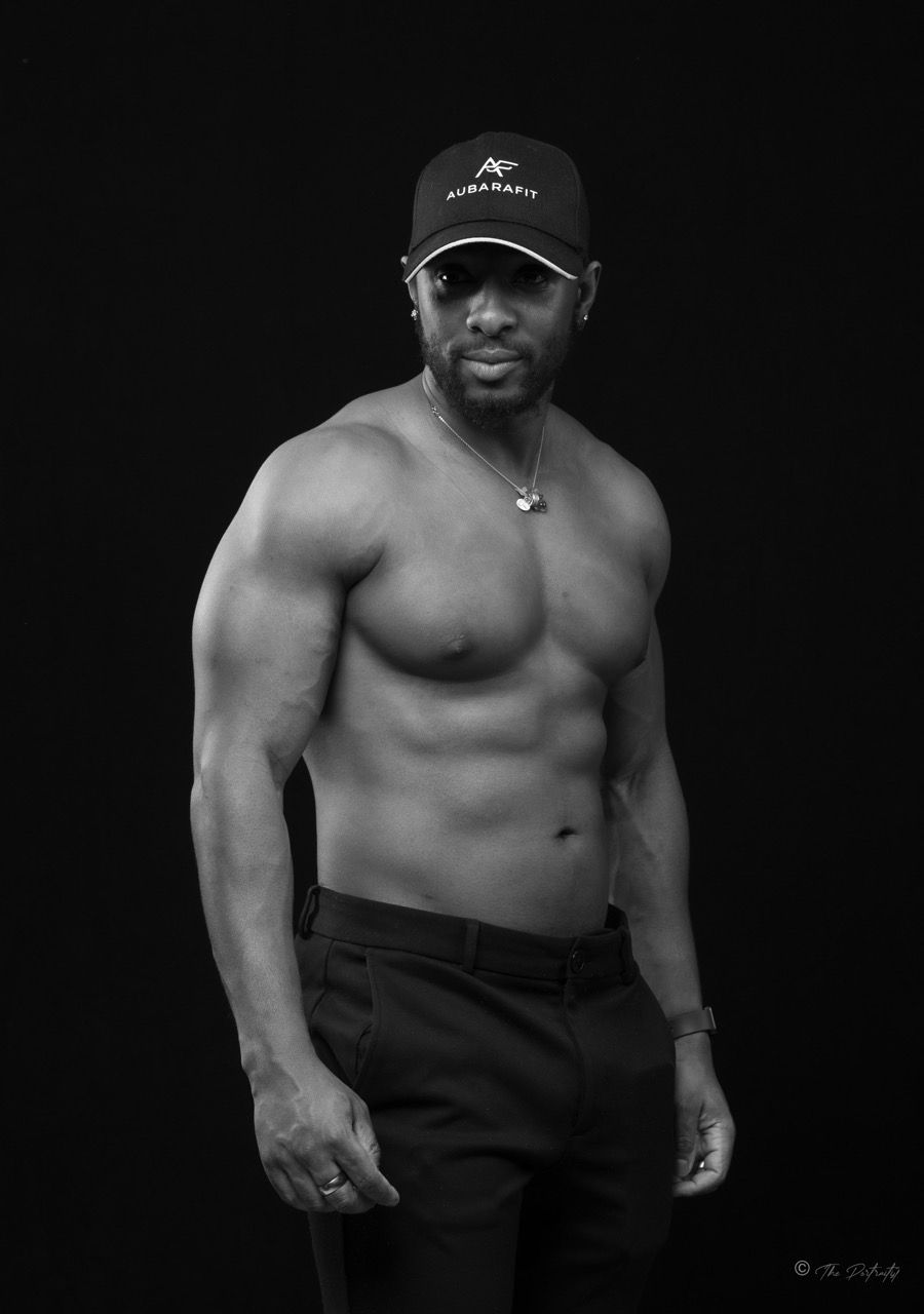 Shirtless muscular man wearing black cap and pants in dramatic black and white fitness portrait.