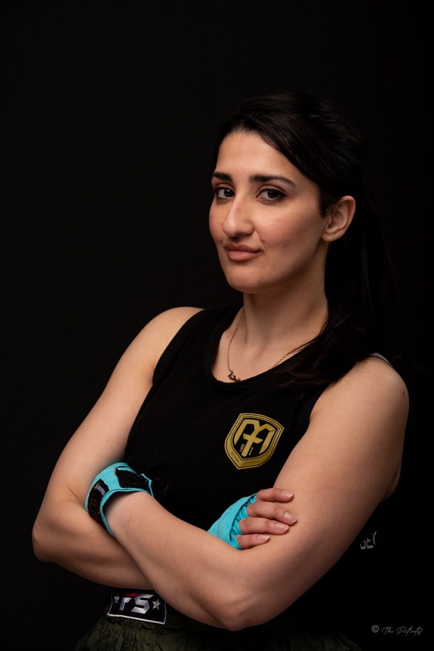 Female boxer with teal hand wraps crossing arms confidently in black sleeveless top against dark background.