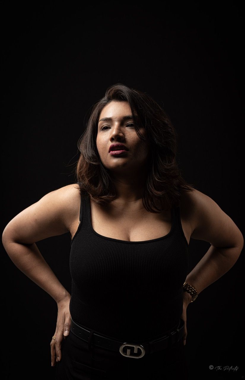 Confident woman in black tank top and belt posing with hands on hips against dark background.