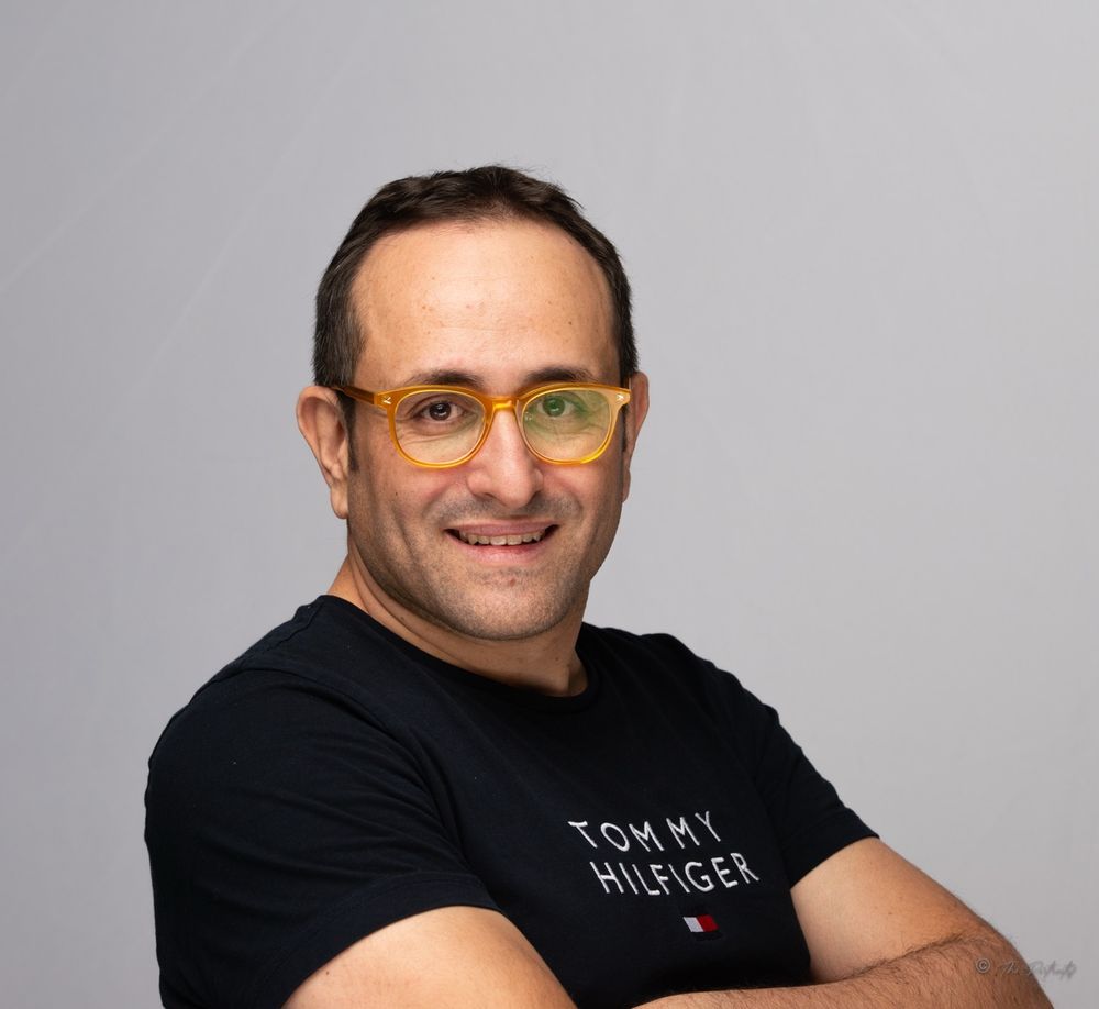Smiling man with orange glasses wearing Tommy Hilfiger black t-shirt with arms crossed on gray background.