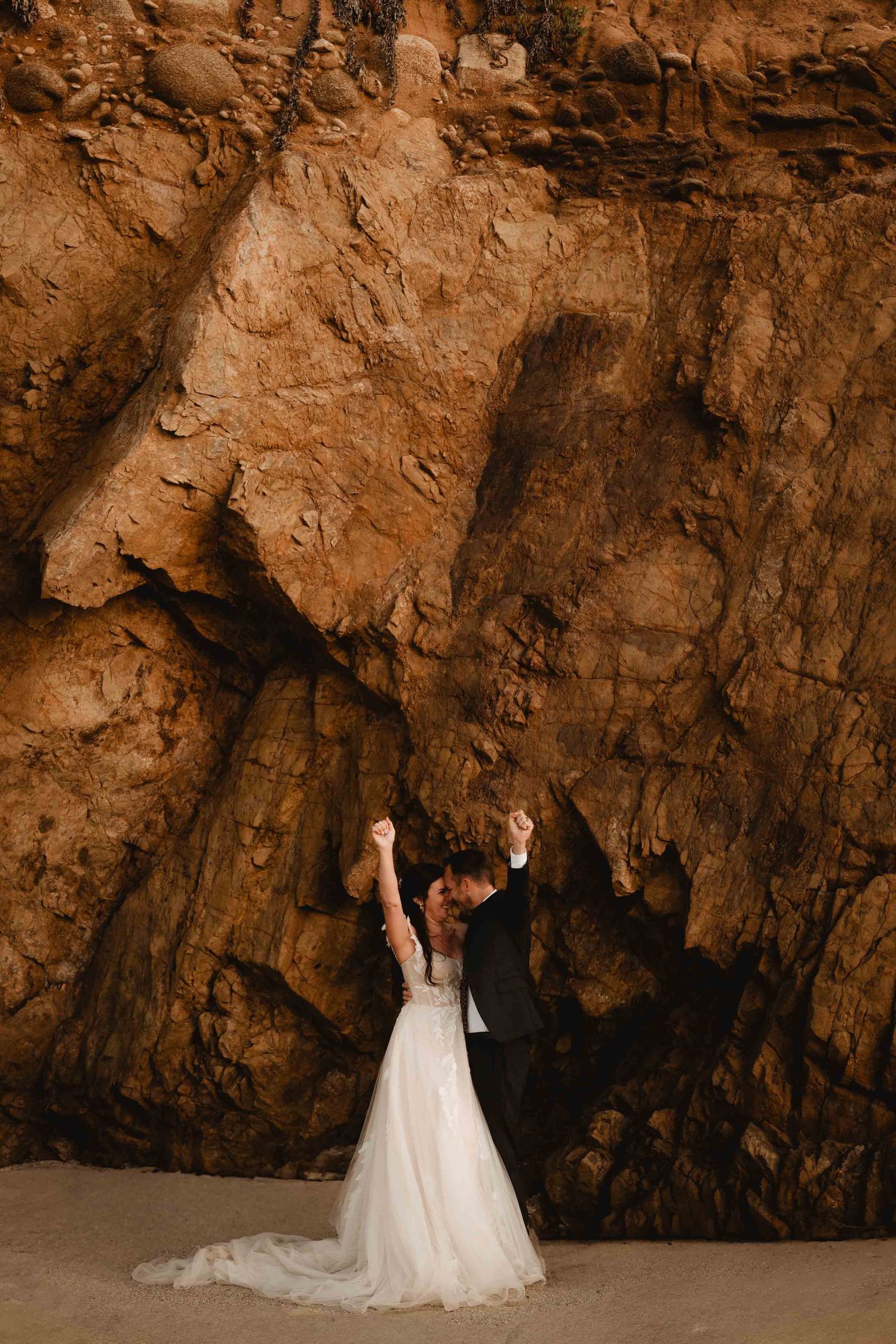 bride and groom holding their hands up victoriously in front of large rock wall