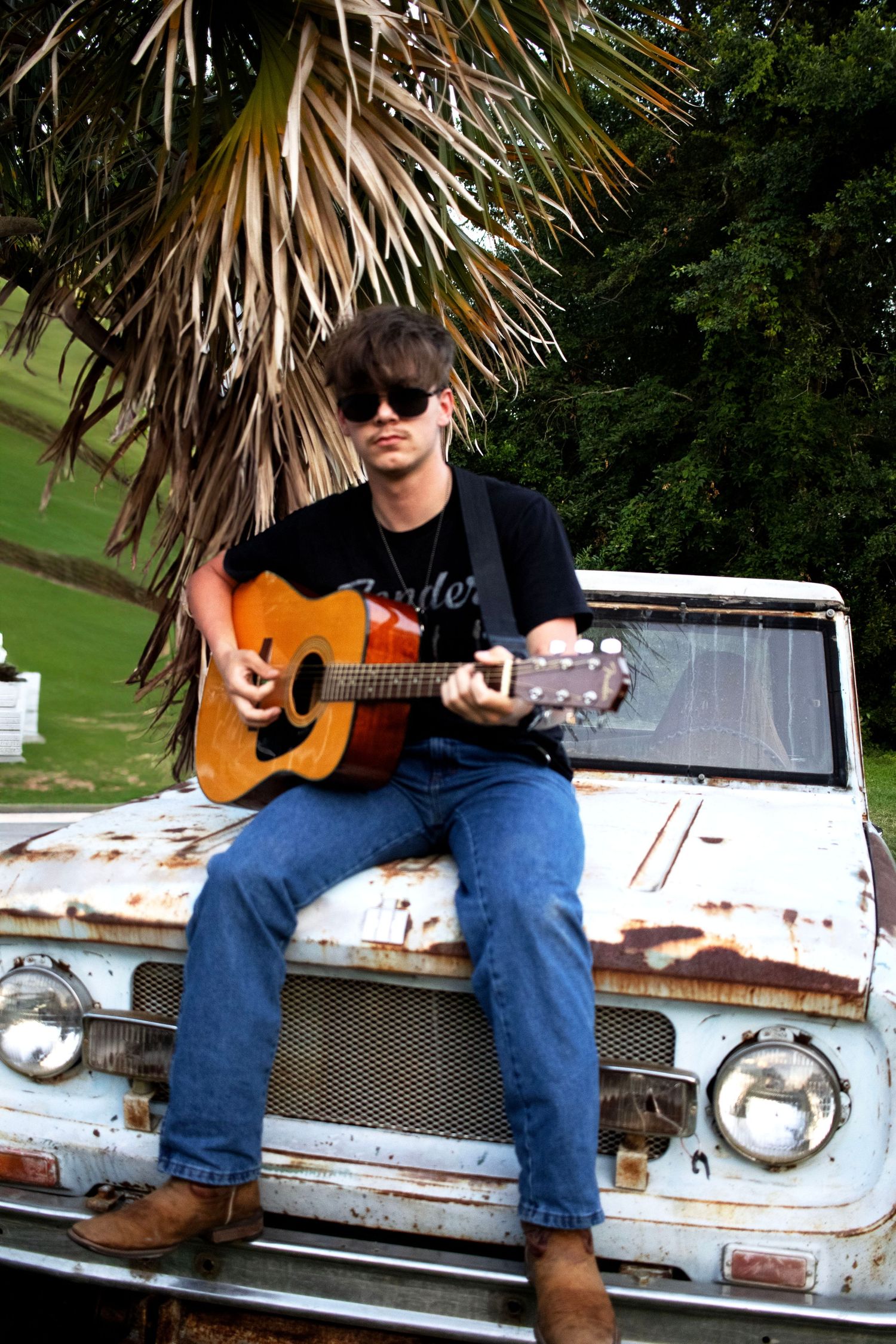 Musician playing acoustic guitar on hood of weathered vintage truck.