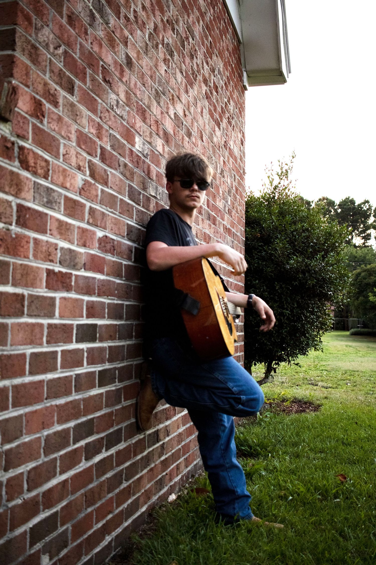 Guitarist leaning against brick wall while playing acoustic instrument outdoors.