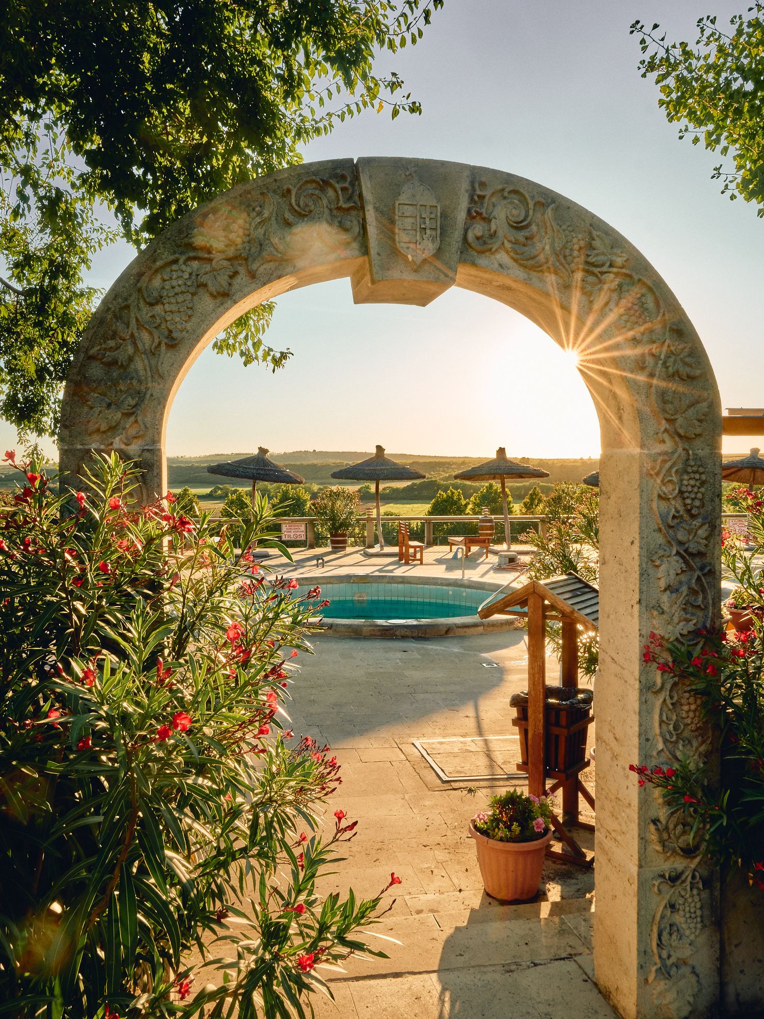 View of thatched roof over red-tiled buildings and swimming pool with rolling countryside hills in background at sunset.